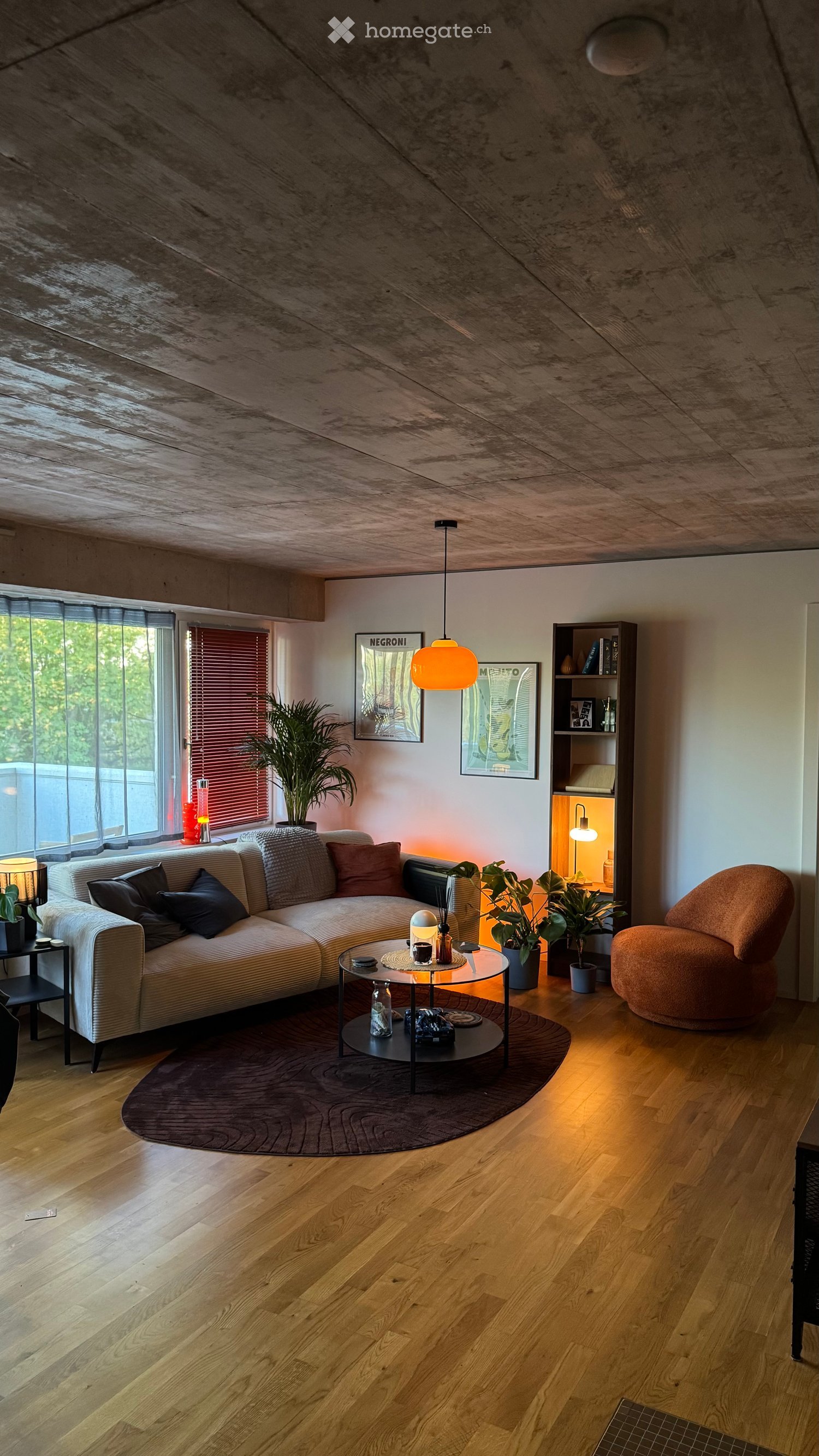 Modern living room with wooden flooring, a sectional sofa, a round glass coffee table, a side table, a large potted plant, a bookshelf, and a hanging lamp.