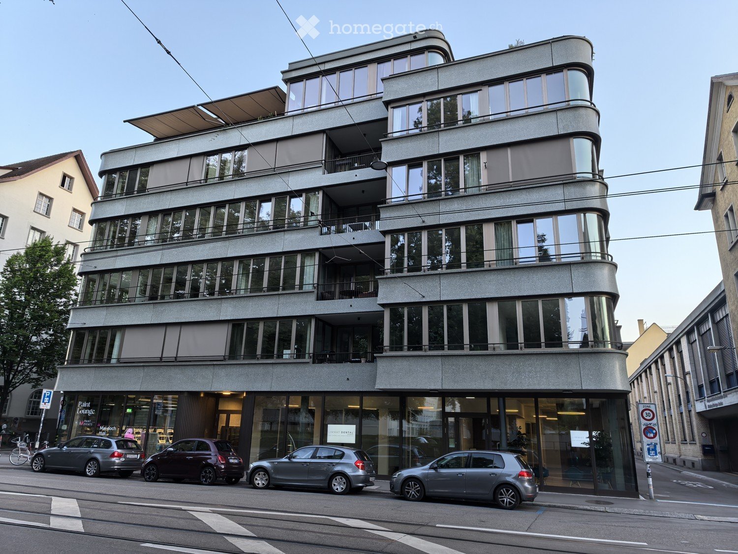 Modern multi-story building with large windows and balconies, located on a street with parked cars and a bike lane.