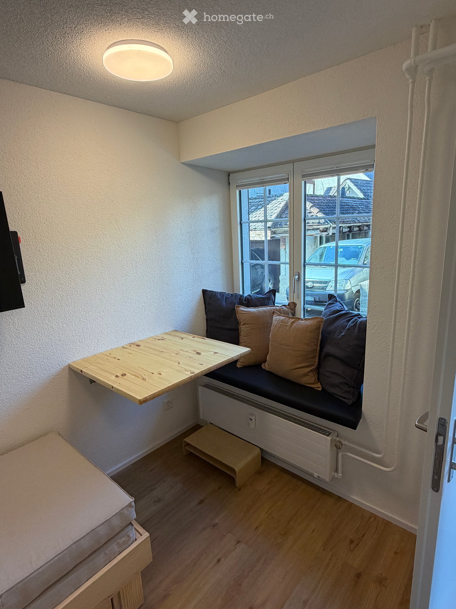 Single bed, wooden floor, foldable table on wall, radiator, window seat with cushions, ceiling lamp, power outlet, mounted television