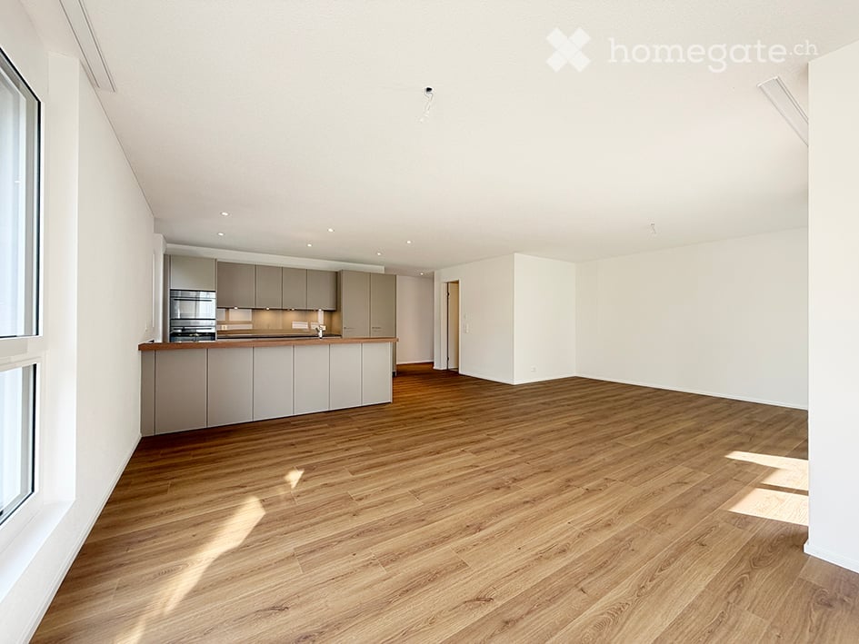 Empty room with wooden flooring, white walls, and a small kitchen area with cabinets and a countertop.