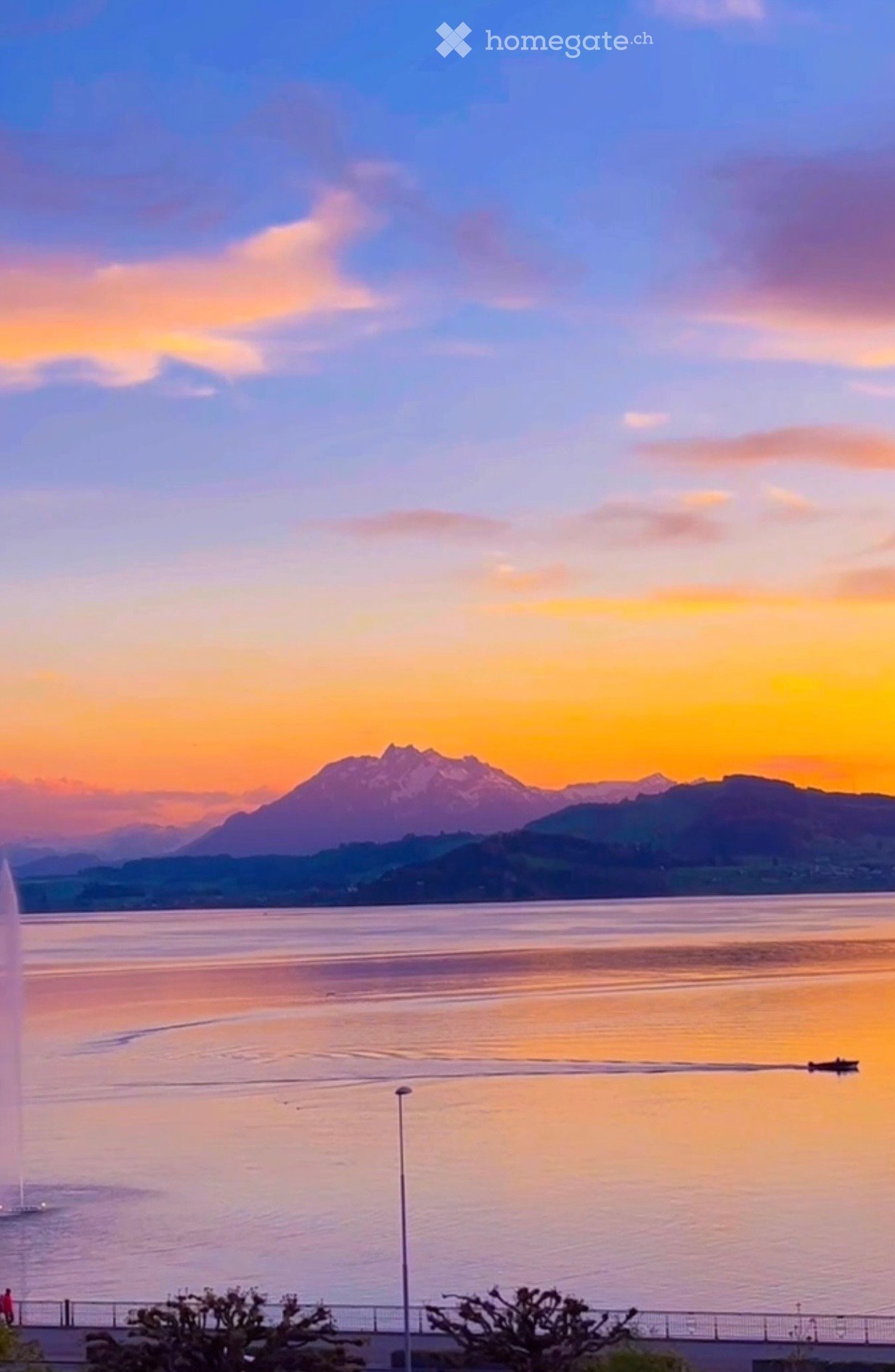 A scenic view of a lake at sunset with a fountain, mountains in the background, and a few people walking along the waterfront.
