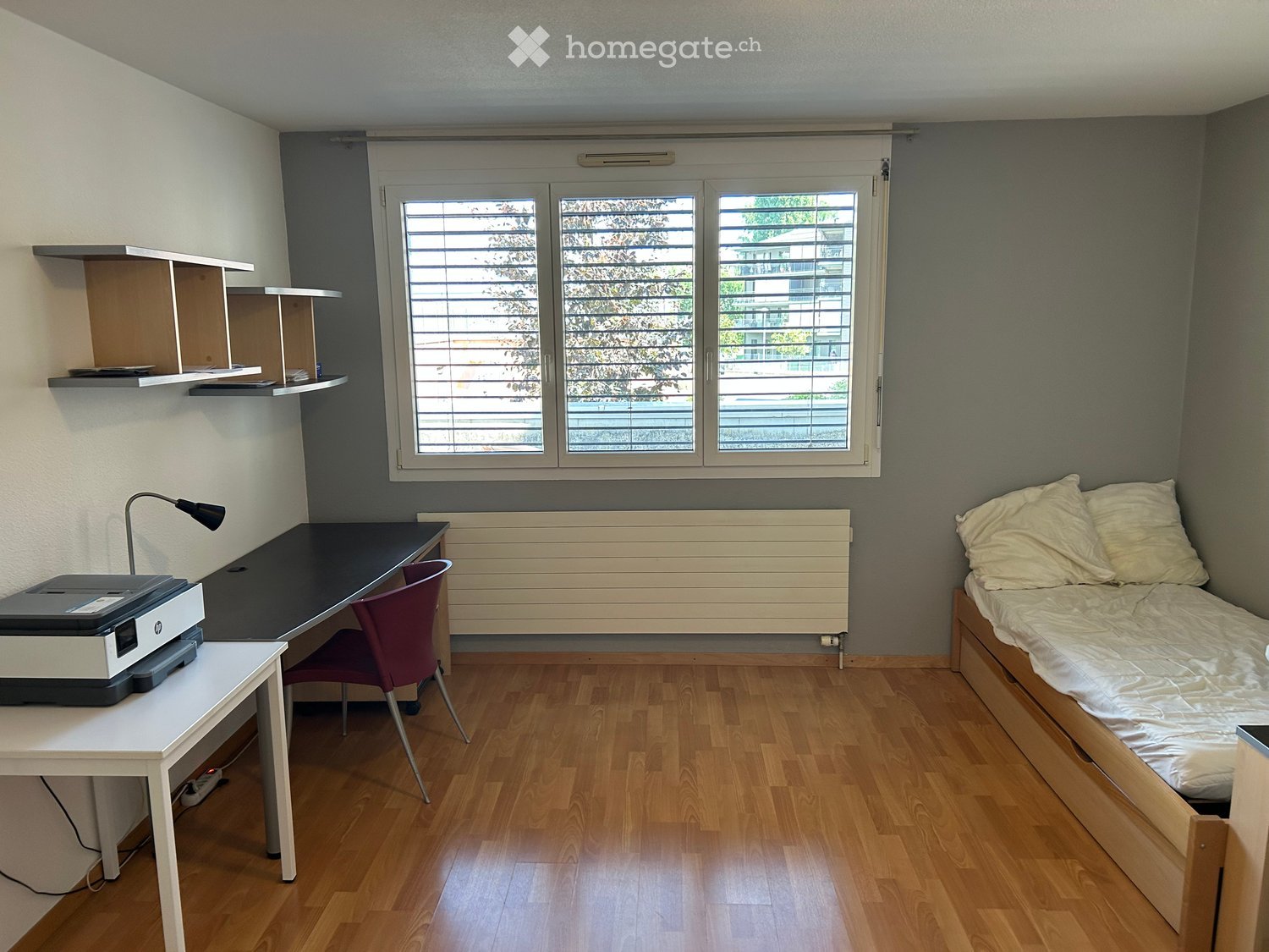 A small, furnished bedroom with a single bed, desk, chair, printer, and shelves. The room features wooden flooring and large windows with blinds.