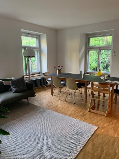 Open-plan living and dining area with wooden flooring, large windows, grey sofa, coffee table, dining table with chairs, floor lamp, and decorative vase.