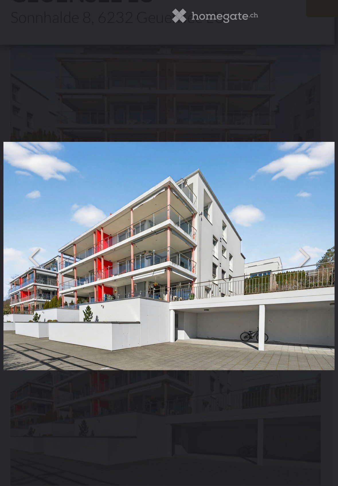 Modern multi-story residential building with white and red accents, balconies, and a paved ground area with a bicycle.