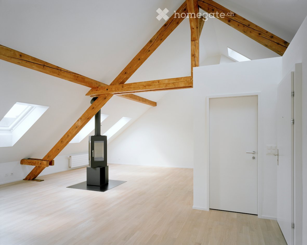Bright, empty attic room with wooden beams, light wooden flooring, white walls, skylights, and a black wood-burning stove.