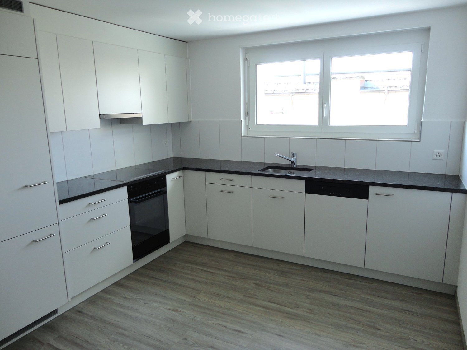 Modern kitchen with white cabinets, black countertops, and stainless steel appliances including a sink, oven, and dishwasher. The space features wooden flooring and large windows.