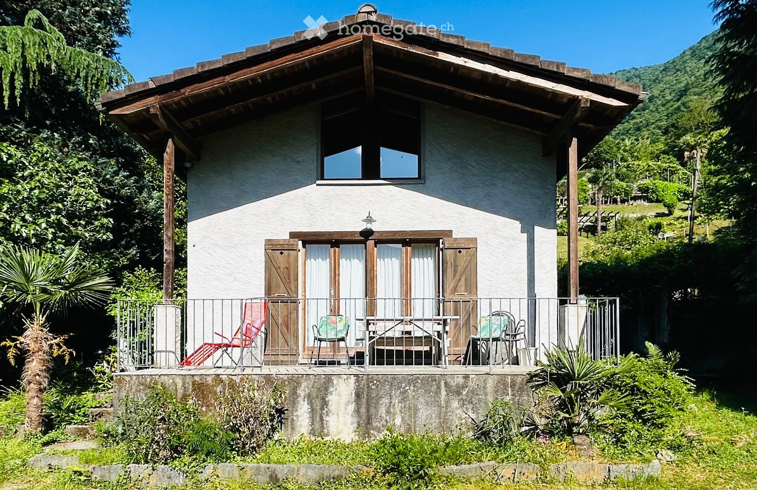 A two-story house with a wooden balcony, surrounded by greenery and a scenic mountain view. The balcony features outdoor furniture including a table, chairs, and a lounge chair.