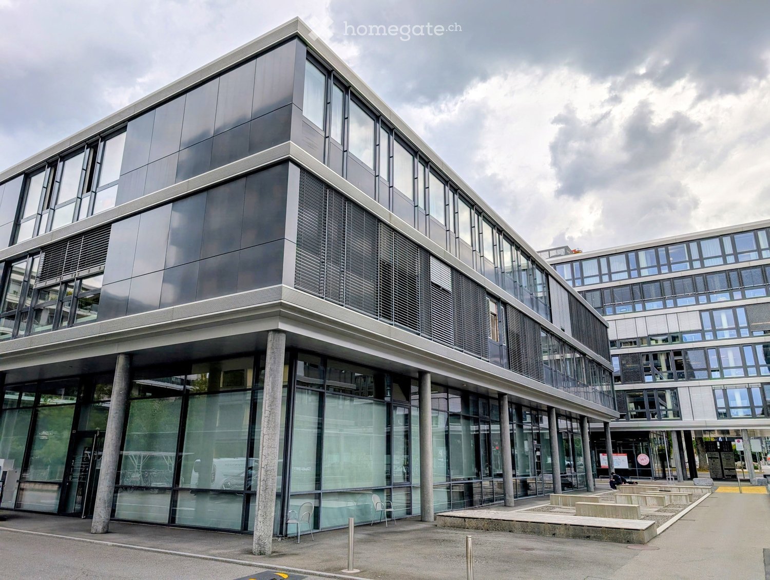 Modern multi-story building with large glass windows and a sleek, contemporary design. Features a raised ground floor with columns and a spacious entrance area.