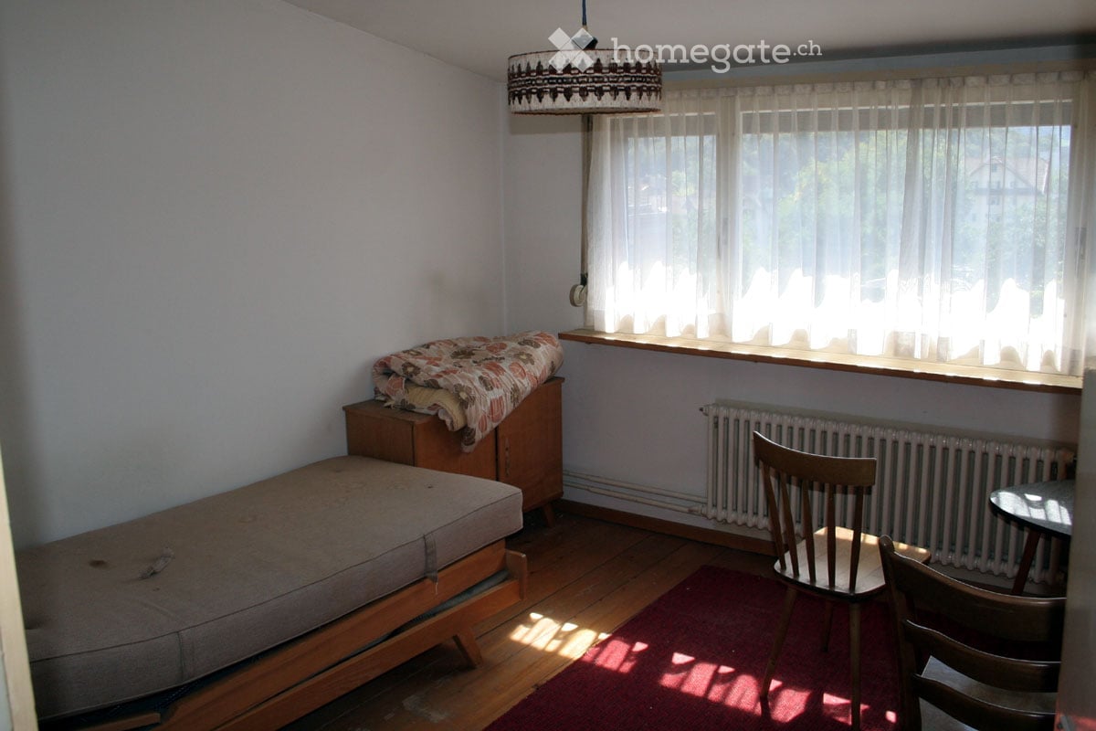 A small, cozy room with wooden flooring, a sofa, a chair, a small table, a radiator, and a window with curtains. A patterned blanket is placed on a wooden cabinet.