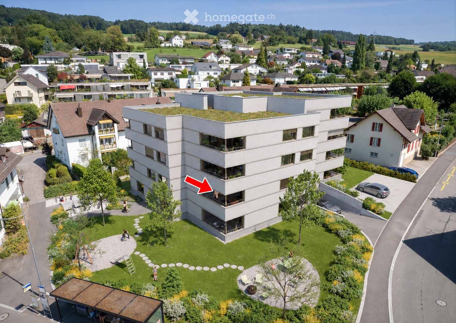 Aerial view of a modern multi-story building with a green roof, surrounded by a landscaped garden area with pathways, trees, and a small seating area. The building is part of a residential neighborhood with other houses and greenery in the background.