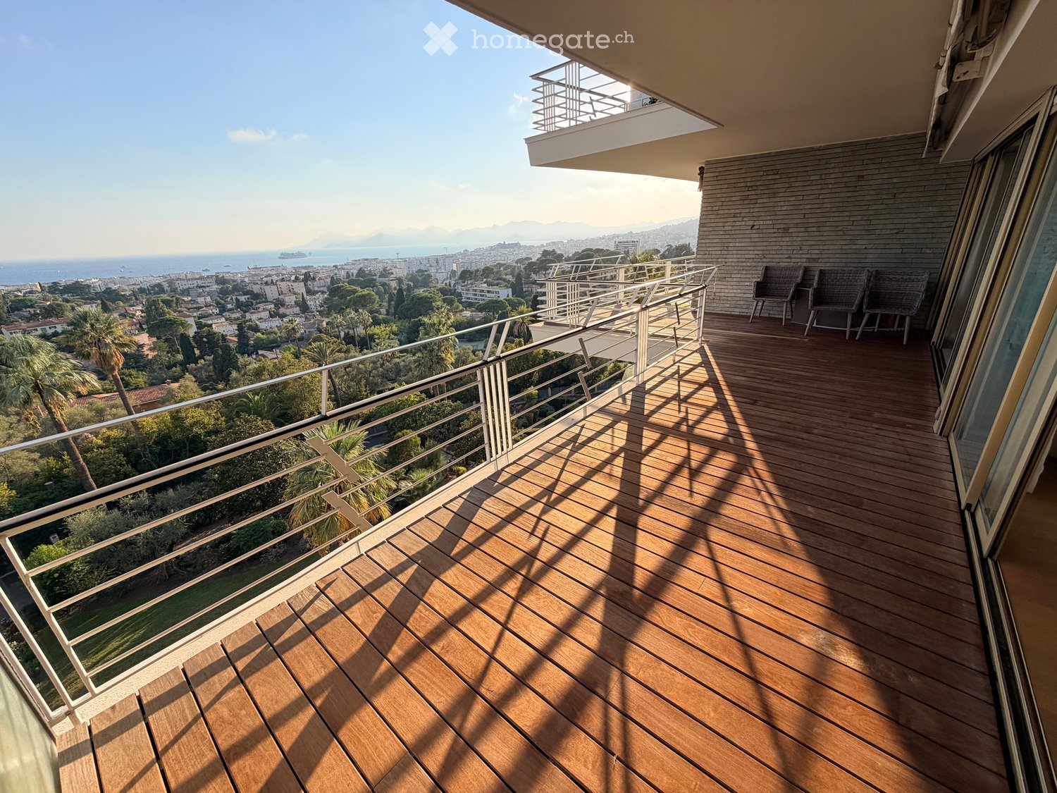 Wooden decked balcony with metal railings, overlooking a scenic coastal cityscape with the sea and distant mountains.