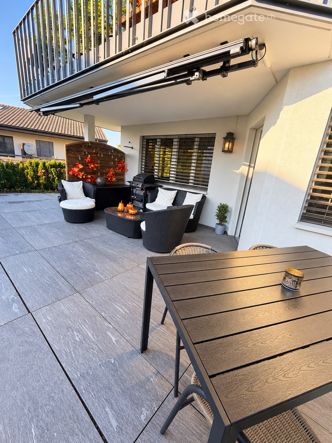 A modern outdoor patio with a wooden dining table, chairs, a wicker sofa set, a coffee table, a grill, and a small potted plant. The area features a tiled floor and is partially covered by a retractable awning.