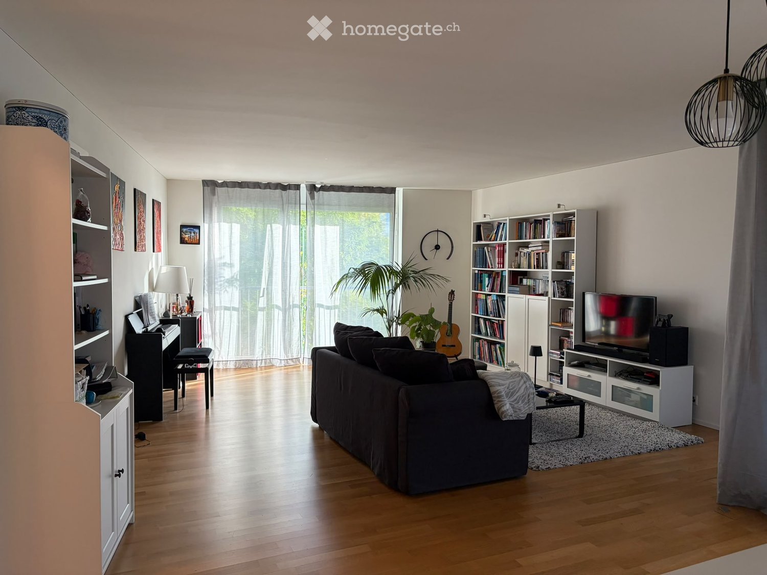 Modern living room with wooden flooring, large windows, a dark-colored sofa, coffee table, bookshelf, piano, and a television stand.