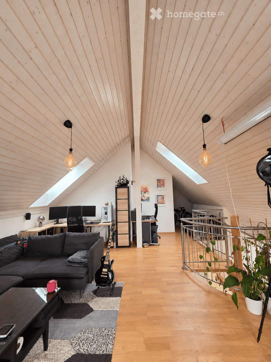 Modern living room with wooden floors, slanted wooden ceiling, large windows, sofa, coffee table, guitar, chair, table, laptop, screen, plant, mirror, and picture frames.