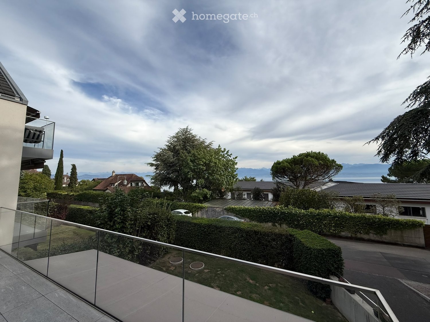 Balcony with glass railing overlooking a residential area, trees, and distant water body under a partly cloudy sky.