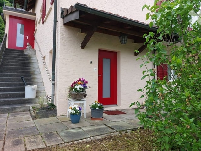 Two-story house with a raised ground floor, red doors, and a small porch area with flower arrangements.