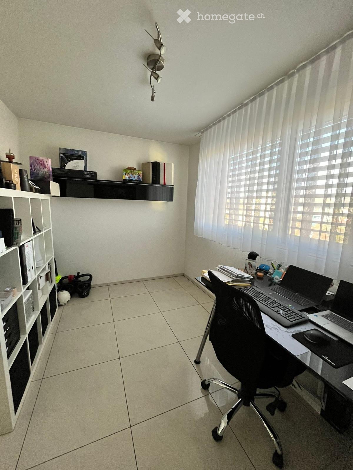 A well-lit office space with a desk, two laptops, a chair, a shelf with books and decorative items, and a window with white curtains.