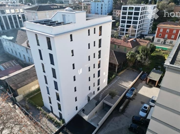 A modern, multi-story residential building with a flat roof and numerous windows, surrounded by other buildings and a parking area.
