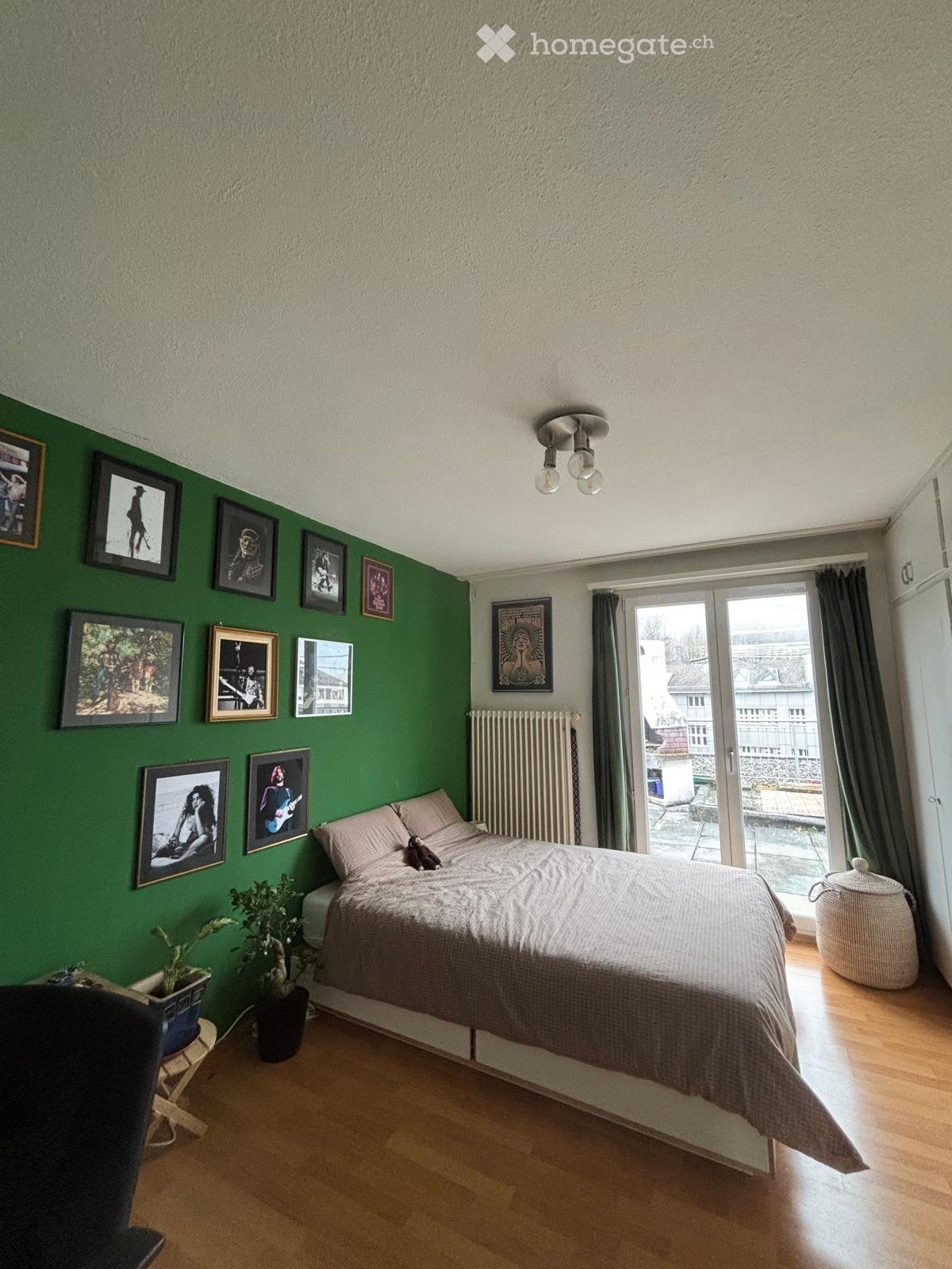 Bedroom with green accent wall, wooden floor, bed with beige bedding, framed pictures, potted plants, and a sliding glass door leading to a balcony.