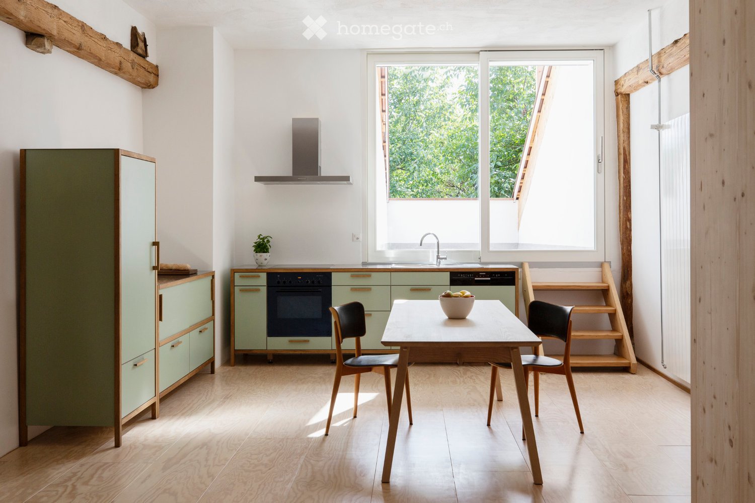 Bright kitchen with light green cabinets, wooden flooring, and a central dining table with chairs. Features an oven, sink, and a small plant on the counter.