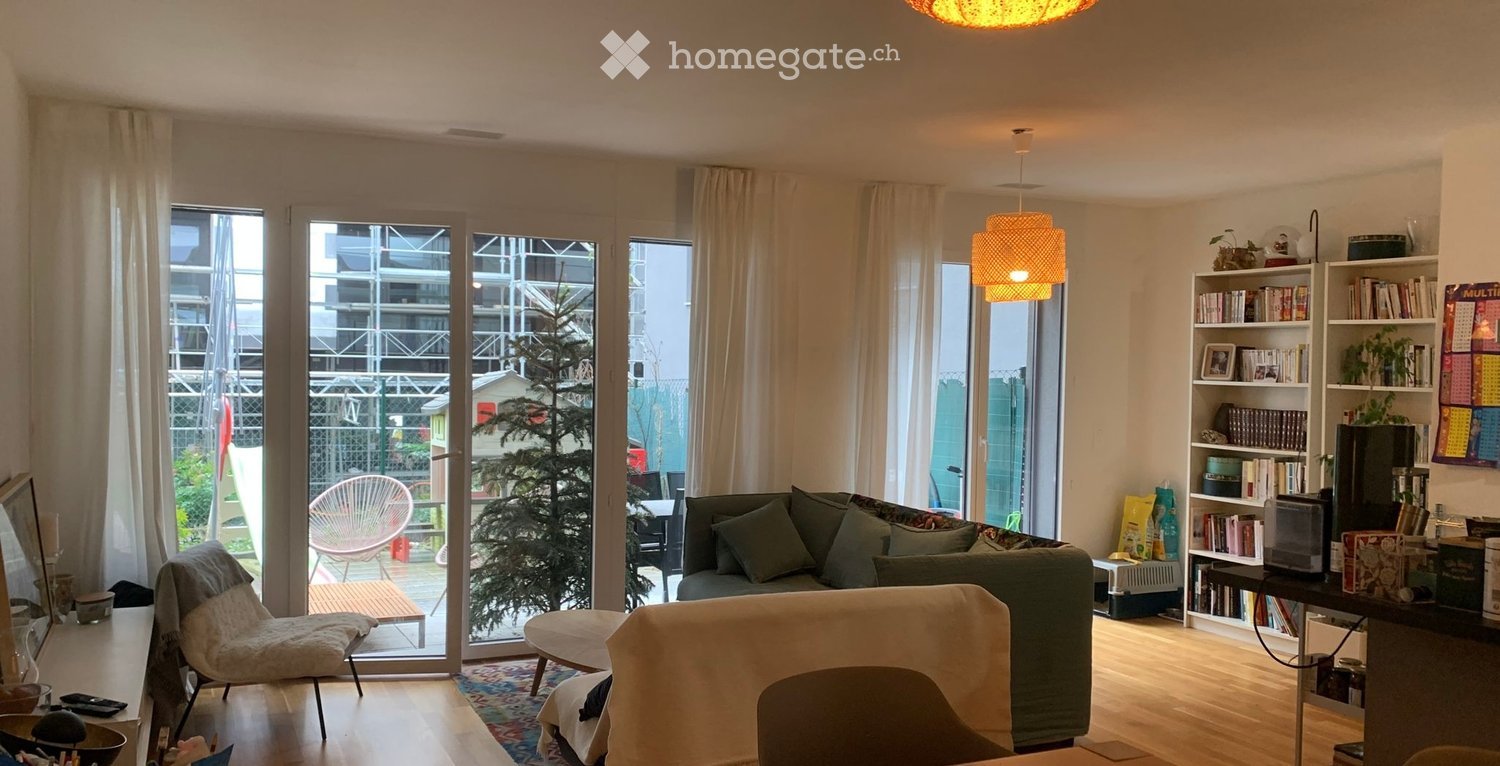 Bright living room with large windows and sliding glass doors leading to a balcony. Features a green sofa, a white chair, a small table, a bookshelf, and a hanging lamp.