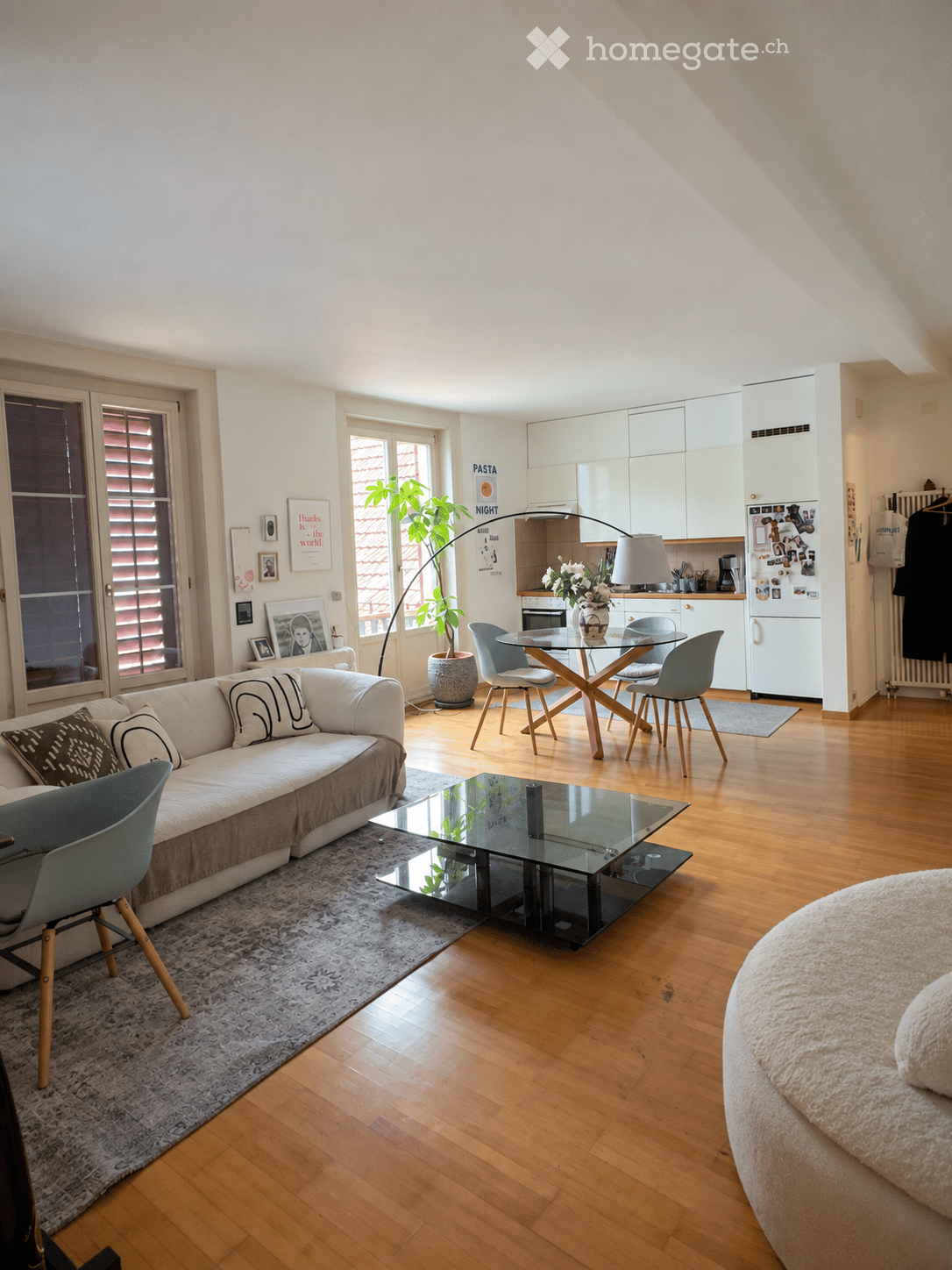 Modern living room with a white sofa, glass coffee table, dining table with chairs, kitchen area with white cabinets, refrigerator, and a potted plant. The room features wooden flooring and large windows with blinds.