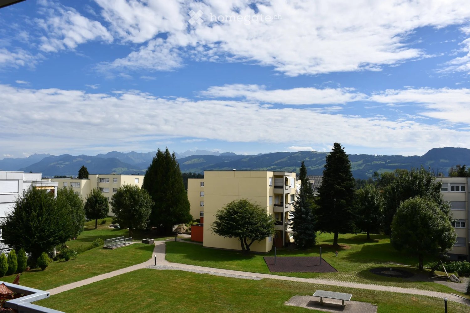 A well-maintained garden area with green lawns, trees, pathways, and buildings in the background, with a scenic mountain view.