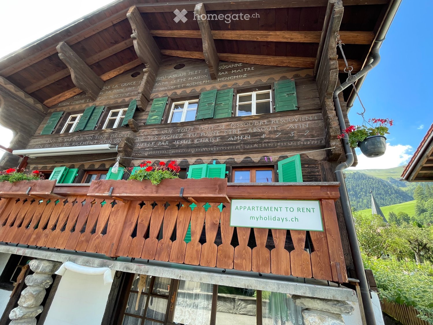 Traditional wooden chalet with green shutters, balconies, and a sign advertising rental apartments. The building features intricate wooden carvings and a stone base.