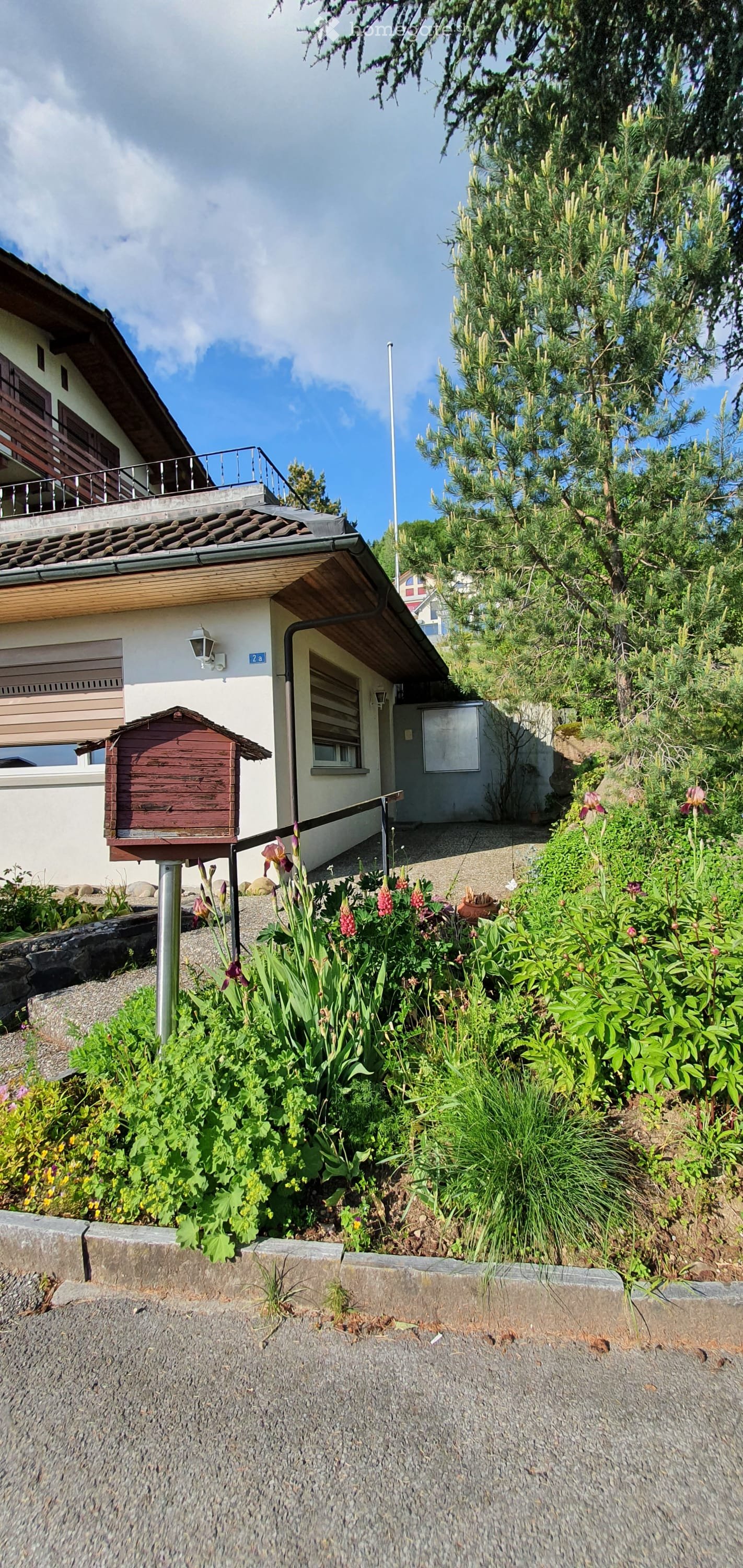 A well-maintained garden area with various plants and flowers, a small wooden structure, and a house with a balcony and garage in the background.