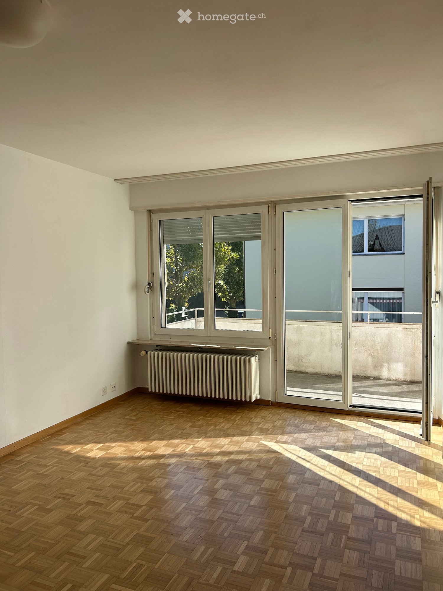 Empty room with wooden herringbone flooring, white walls, large windows with blinds, and a radiator under one window.