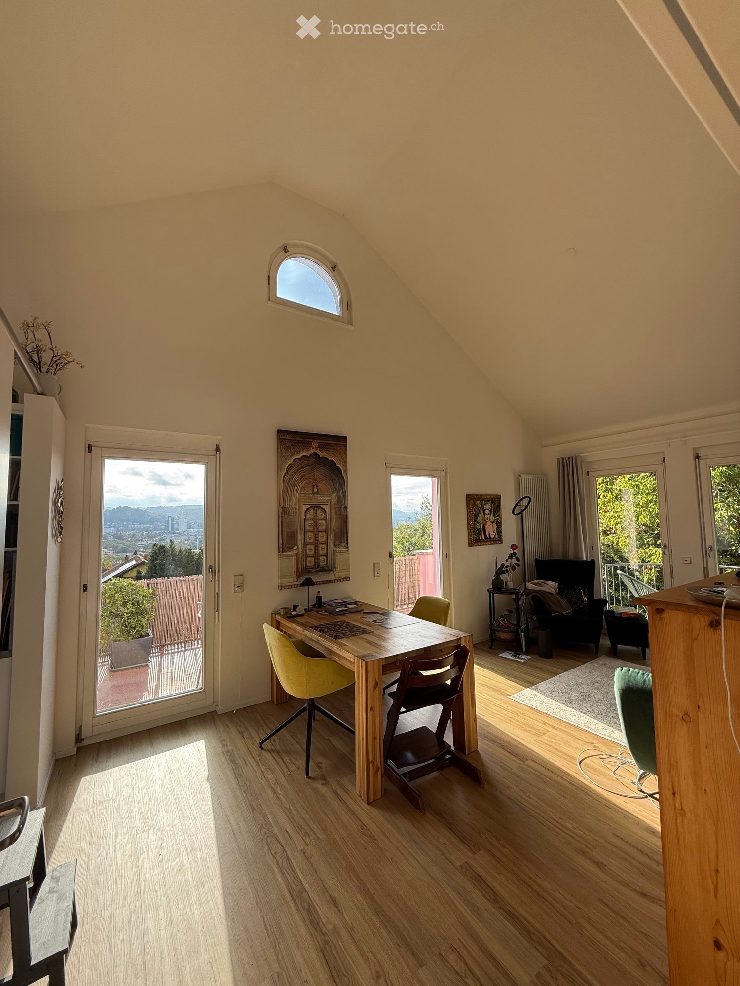 Bright, airy room with wooden flooring, large windows, and a view. Features a wooden desk with chairs, a sofa, a lamp, and a picture frame on the wall.