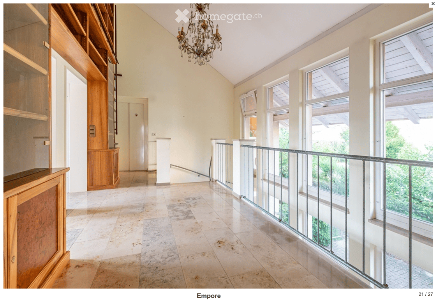 Bright, spacious hallway with high ceilings, large windows, and a chandelier. Features a wooden cabinet on the left and a staircase leading upwards.
