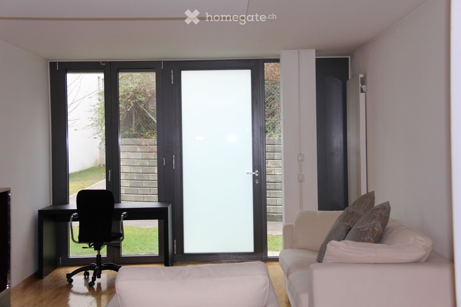 A modern living room with a white sofa, ottoman, desk, and chair, featuring large sliding glass doors leading to an outdoor area.