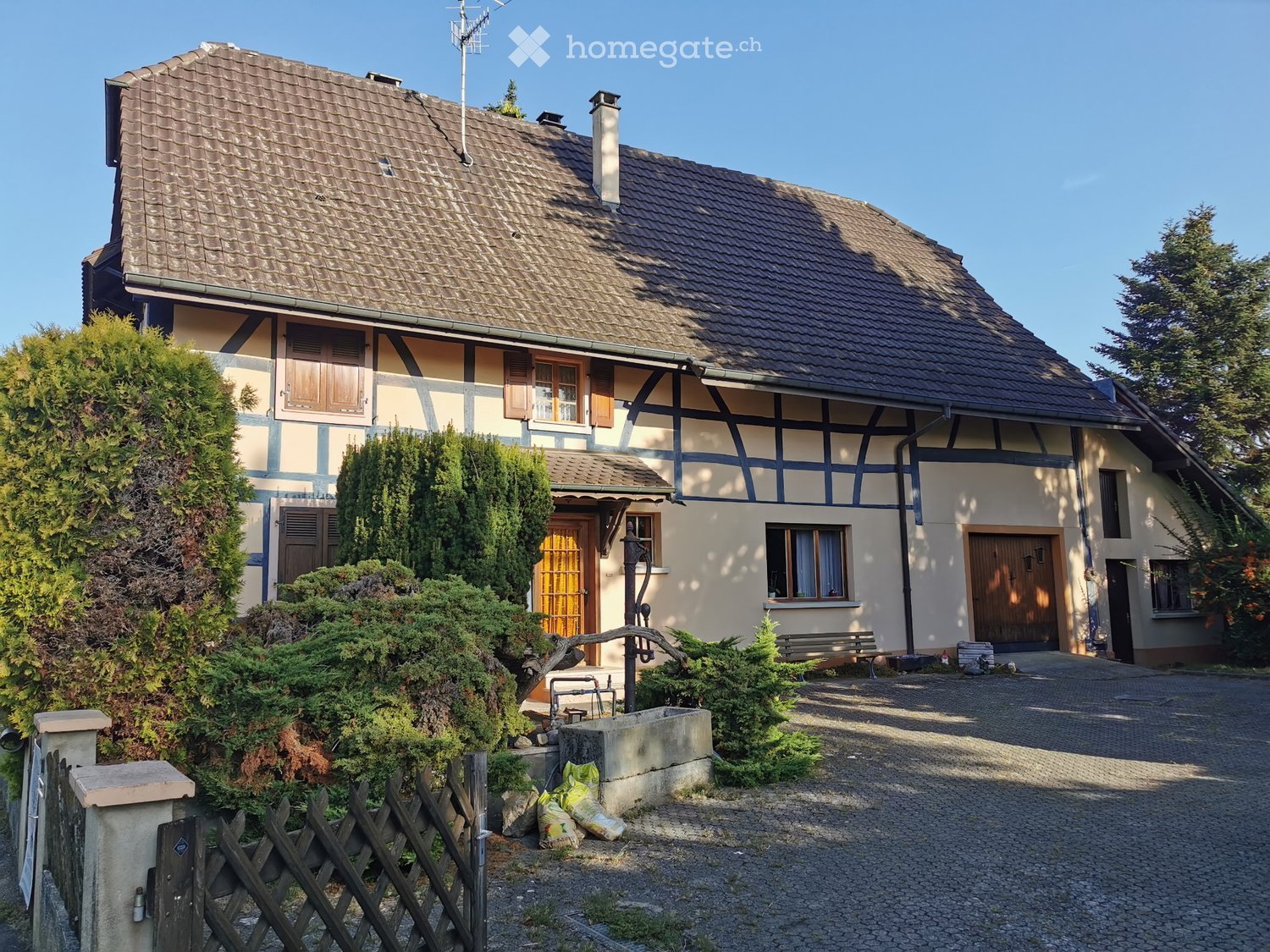 Two-story house with a tiled roof, wooden shutters, and a traditional half-timbered facade. The house features a small garden with a bench, a well, and a wooden fence.