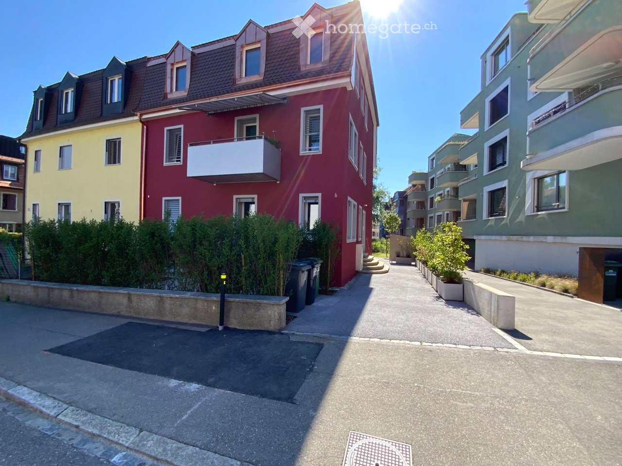 A modern residential building with a red facade, white balcony, and multiple windows. The building is surrounded by greenery and other residential structures.