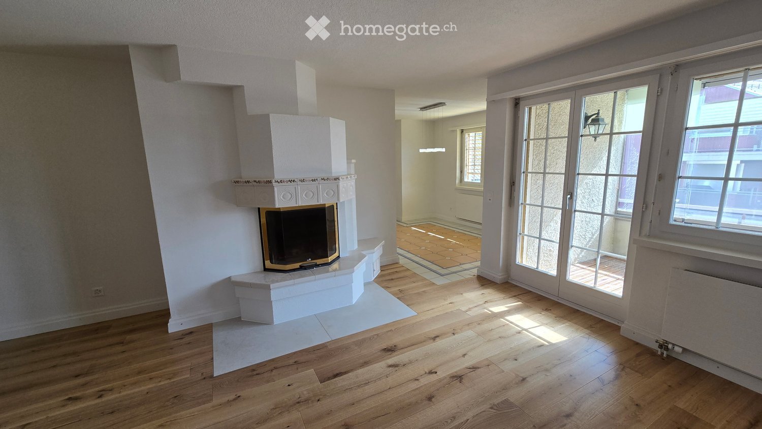 Bright living room with wooden flooring, fireplace, large windows, and glass doors leading to a tiled area.