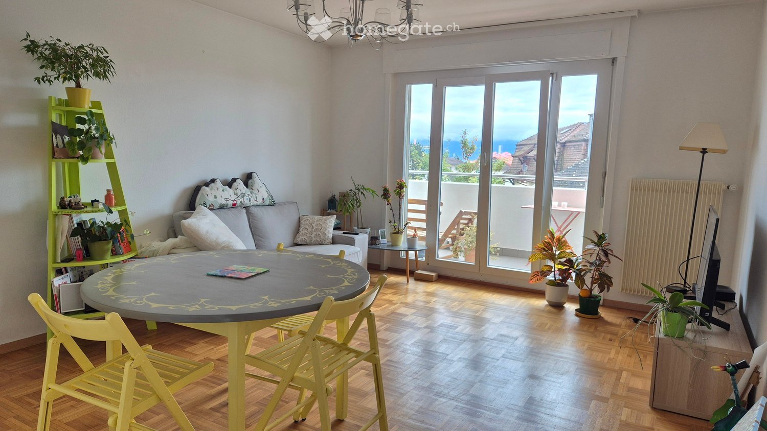 Bright living room with wooden flooring, large windows offering a scenic view, a round table with chairs, a sofa with decorative pillows, a green shelving unit with plants and books, and a chandelier hanging from the ceiling.
