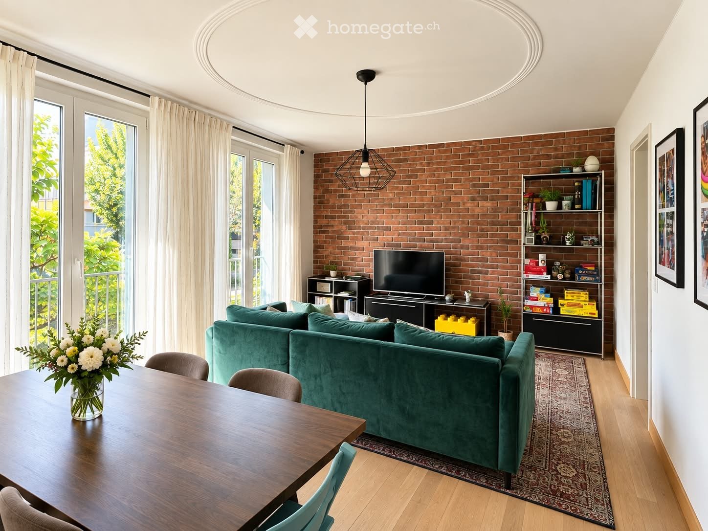 Modern living room with a green sofa, wooden dining table, chairs, television, bookshelf, and decorative items.