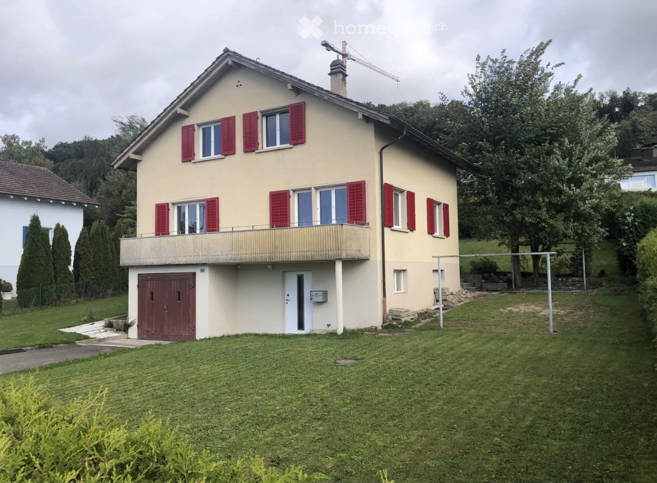 Two-story house with beige walls, red shutters, and a balcony on the upper floor. Features a garage entrance and a well-maintained lawn.