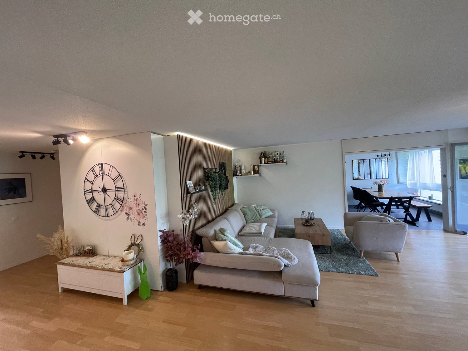 Modern living room with light-colored furniture, a sectional sofa, coffee table, side table, and a dining area visible through an open doorway.