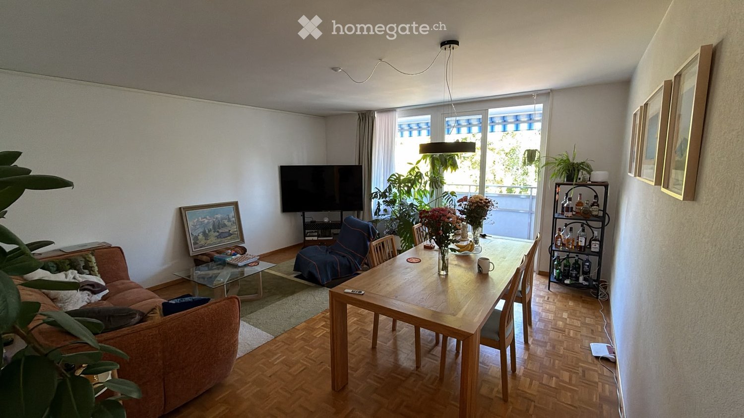 A modern living room with a wooden dining table, sofa, TV, and decorative items, featuring large windows and plants.