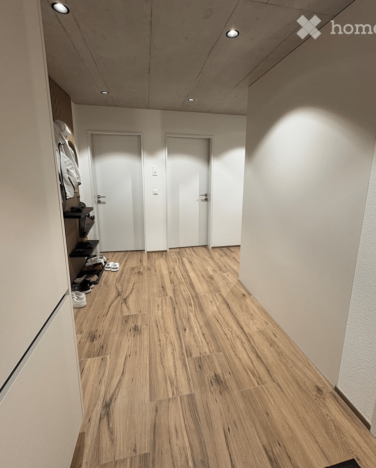 Clean, modern hallway with light wooden flooring, white walls, and recessed ceiling lighting. Two white doors are visible at the end of the hallway, with a built-in shoe rack on the left side holding shoes and jackets.