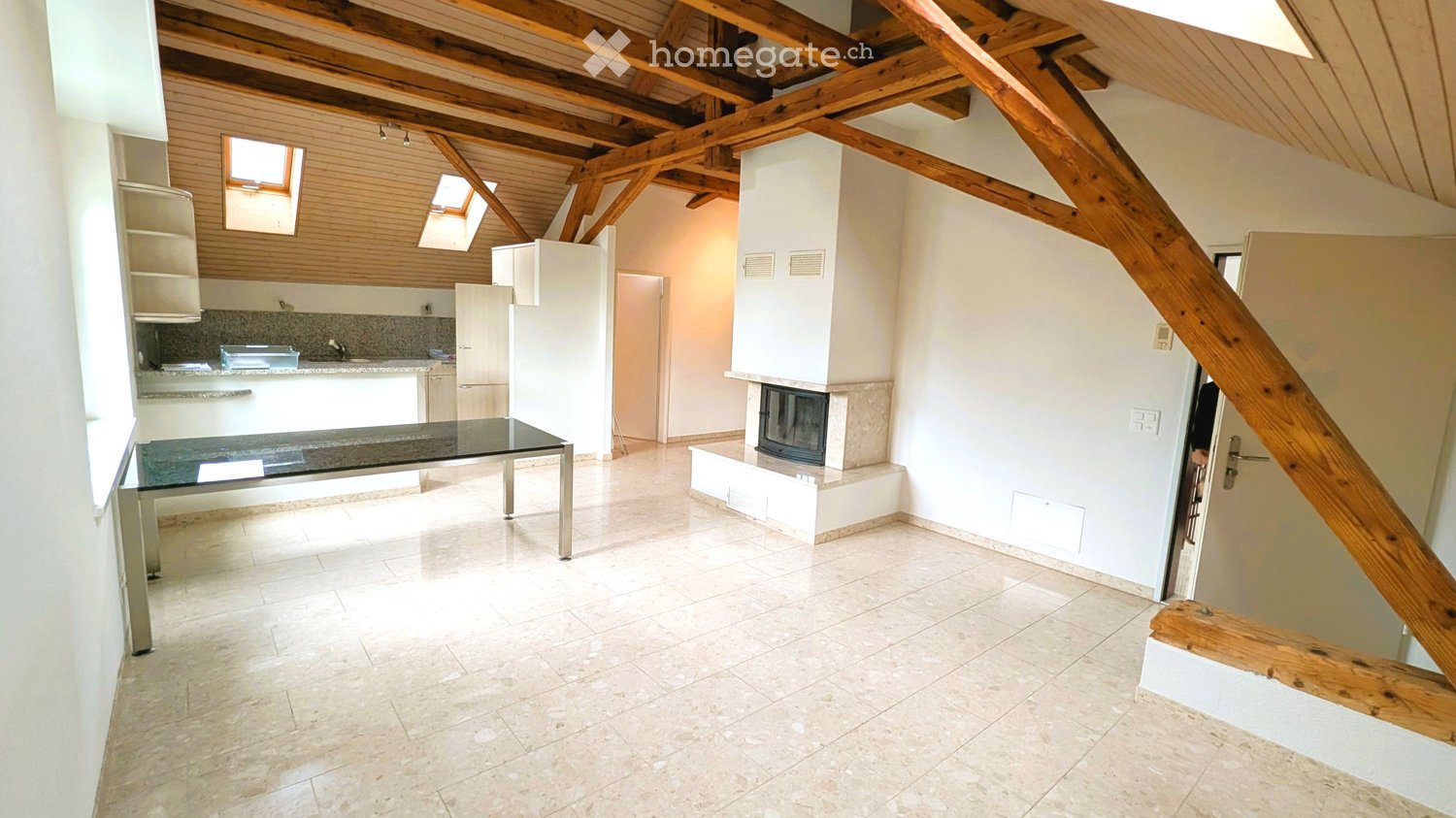 Open living and kitchen area with exposed wooden ceiling beams, white walls, tiled floor, fireplace, glass table, and an open kitchen with countertops and storage units.