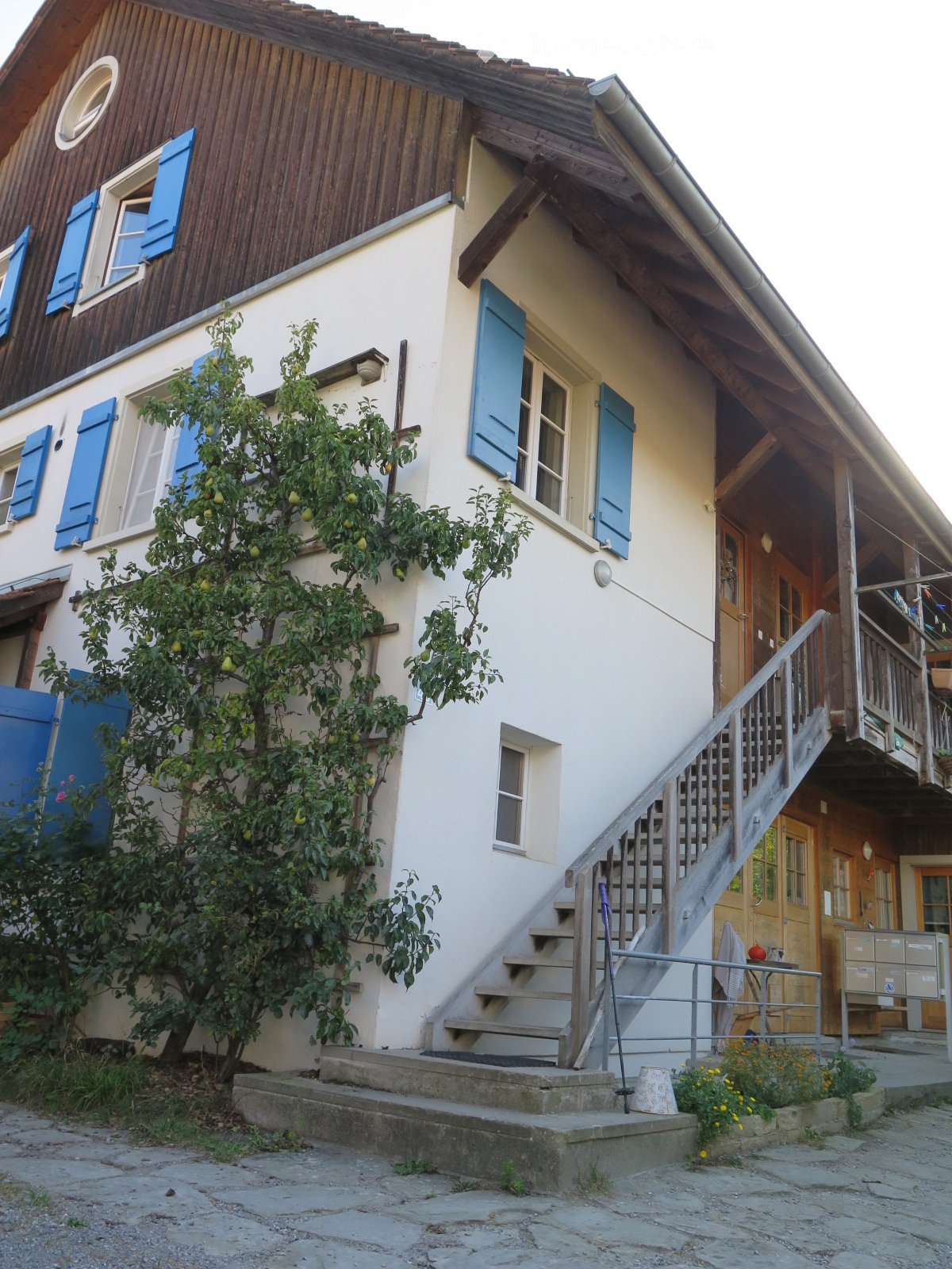 Two-story house with a raised ground floor, wooden exterior, blue shutters, and a staircase leading to the entrance.