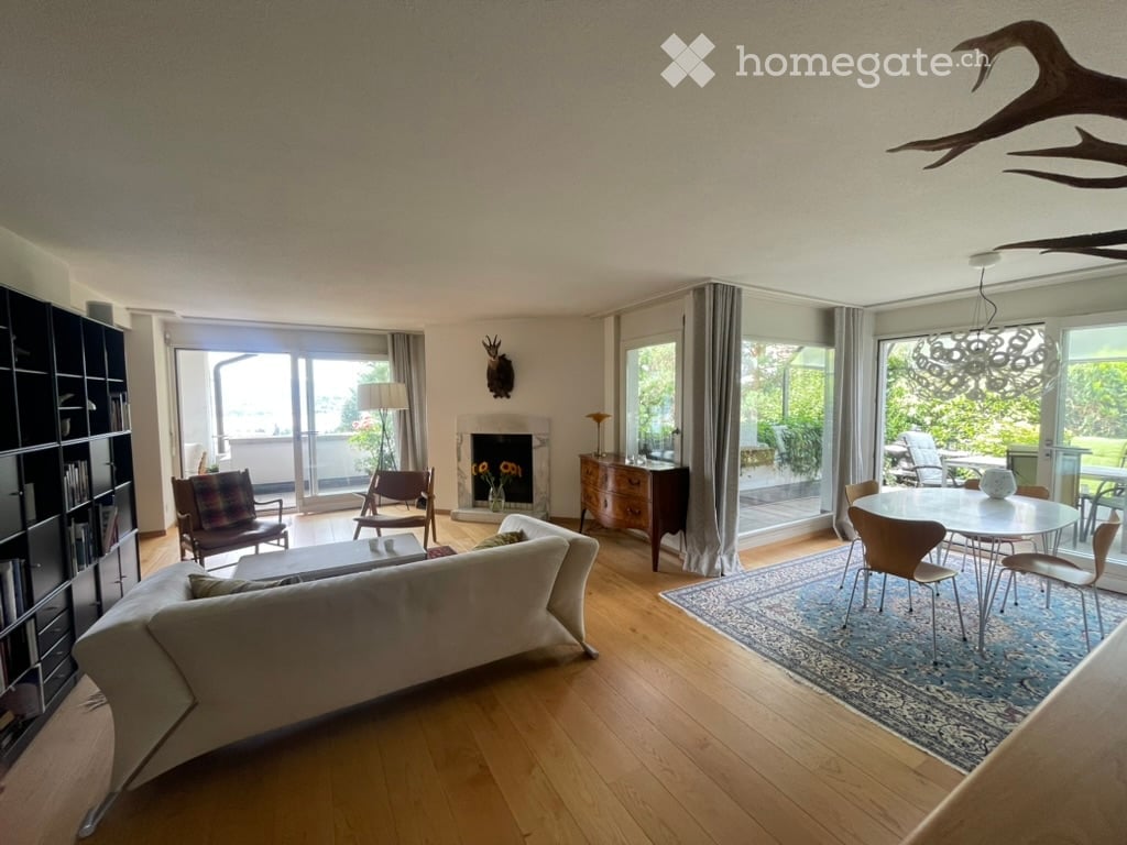 Spacious living room with wooden flooring, large windows offering a view, a fireplace, a sofa, coffee table, armchairs, dining table with chairs, bookshelf, and decorative items.