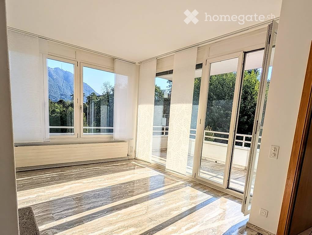 Bright, unfurnished room with large windows and sliding glass doors leading to a balcony, featuring wooden flooring and a scenic mountain view.