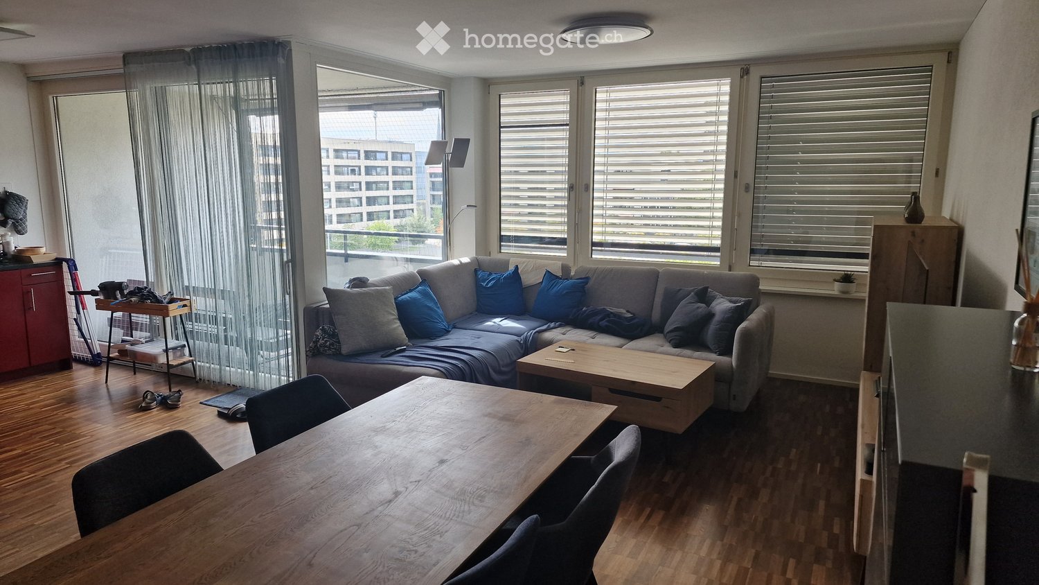 Modern living room with a grey sectional sofa, wooden coffee table, dining table with chairs, and large windows with blinds. The room features a wooden floor and a view of a cityscape.