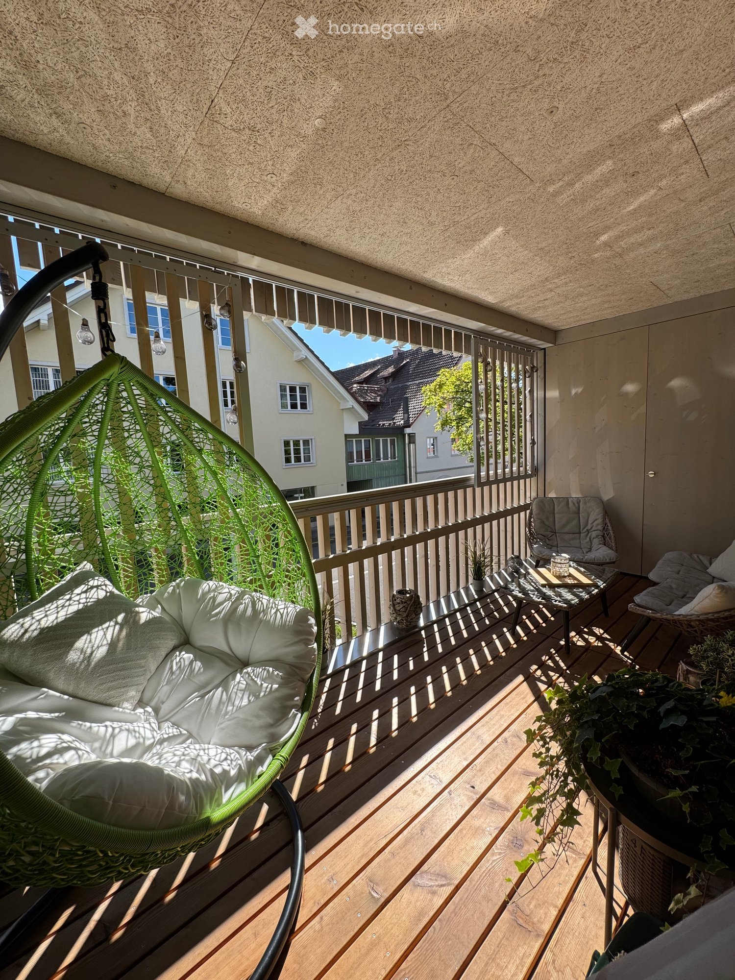 Balcony with wooden decking, a wicker hanging chair, a small table, a chair, and potted plants.