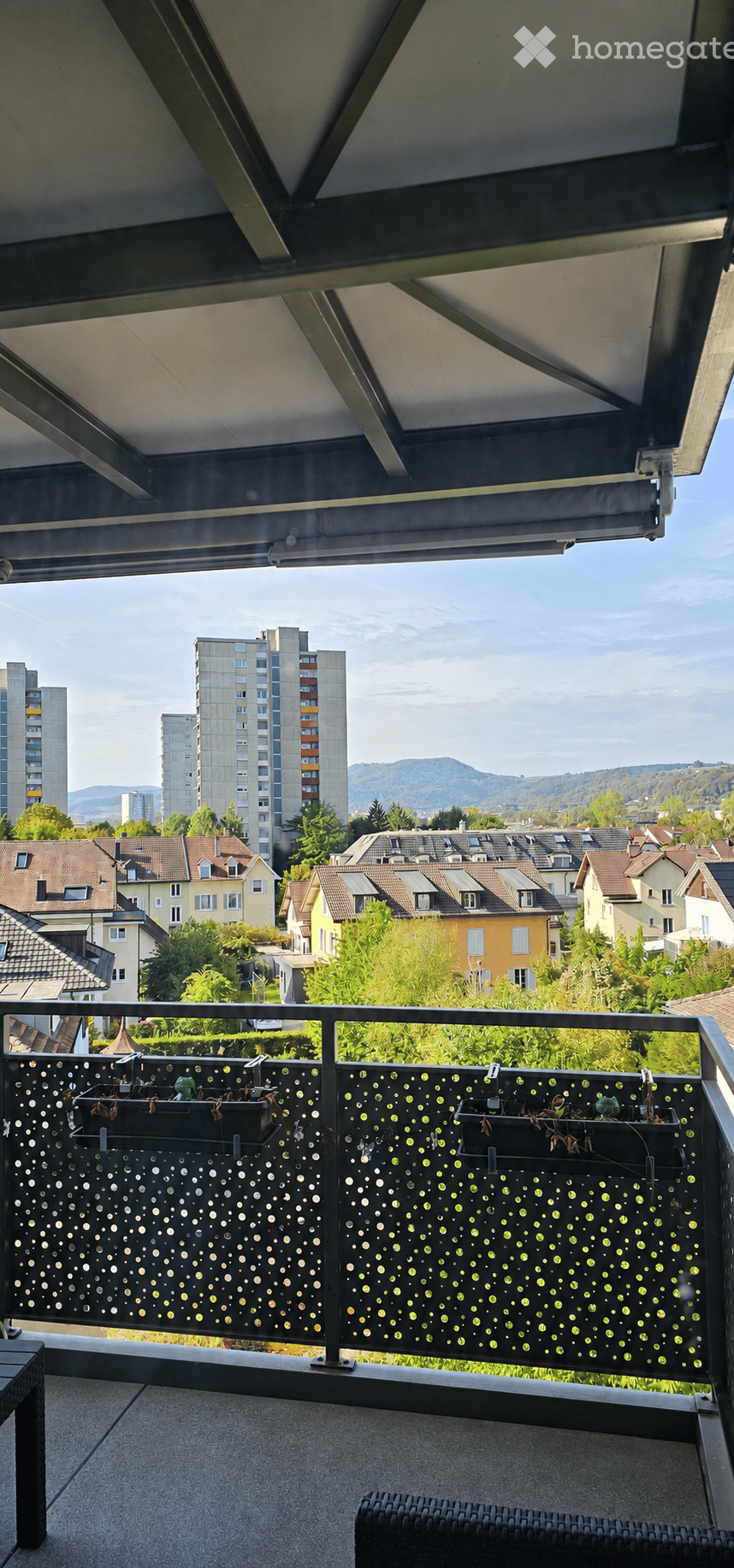 Balcony with modern railing, potted plants, and outdoor furniture overlooking a cityscape with residential buildings and distant mountains.