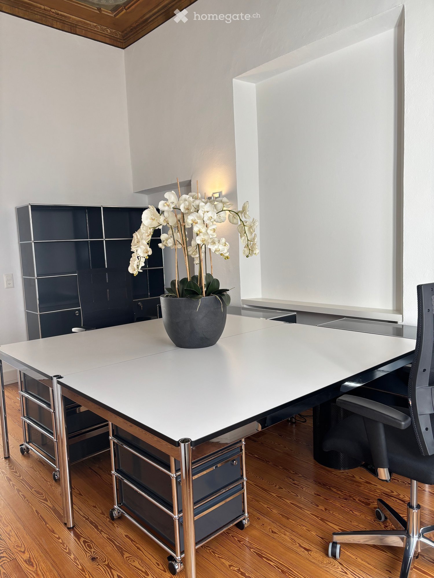 Modern office space with a large white table, black office chair, vase with white orchids, and dark cabinet.